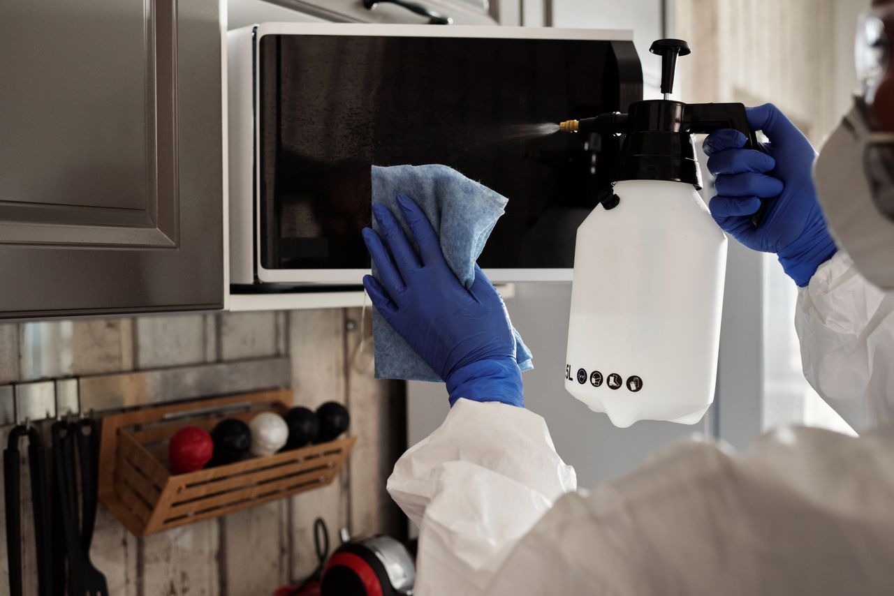 Person wearing protective equipment cleaning a microwave indoors with a spray bottle and cloth.