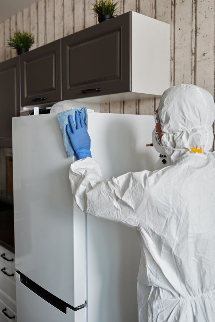 A healthcare professional wearing protective equipment disinfects a refrigerator to help maintain safety and cleanliness.